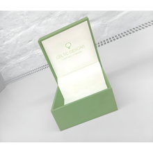 Lade das Bild in den Galerie-Viewer, Geschenkbox für keltischen Schmuck. Grüne Schmuckschatulle mit der Inschrift Celtic Designs, geöffnet  auf einer weißen Platte liegend. Im Hintergrund ist eine weiss  gestrichenes Ziegelwand.

